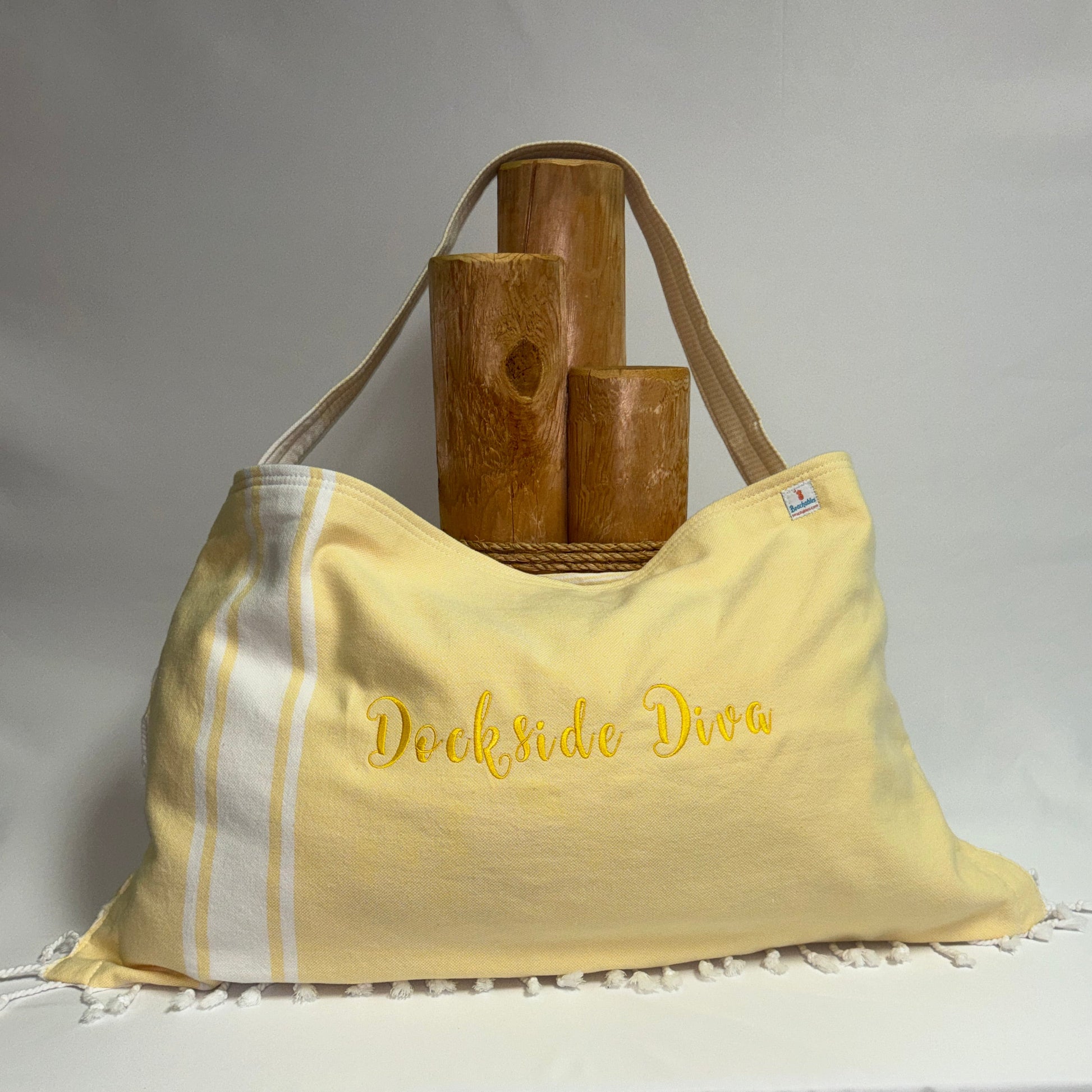 A pale yellow woven tote bag with white stripes and white fringe, featuring the words 'Dockside Dive' embroidered in yellow on the front. The bag is styled with three wooden cylinder props on a draped white fabric background.
