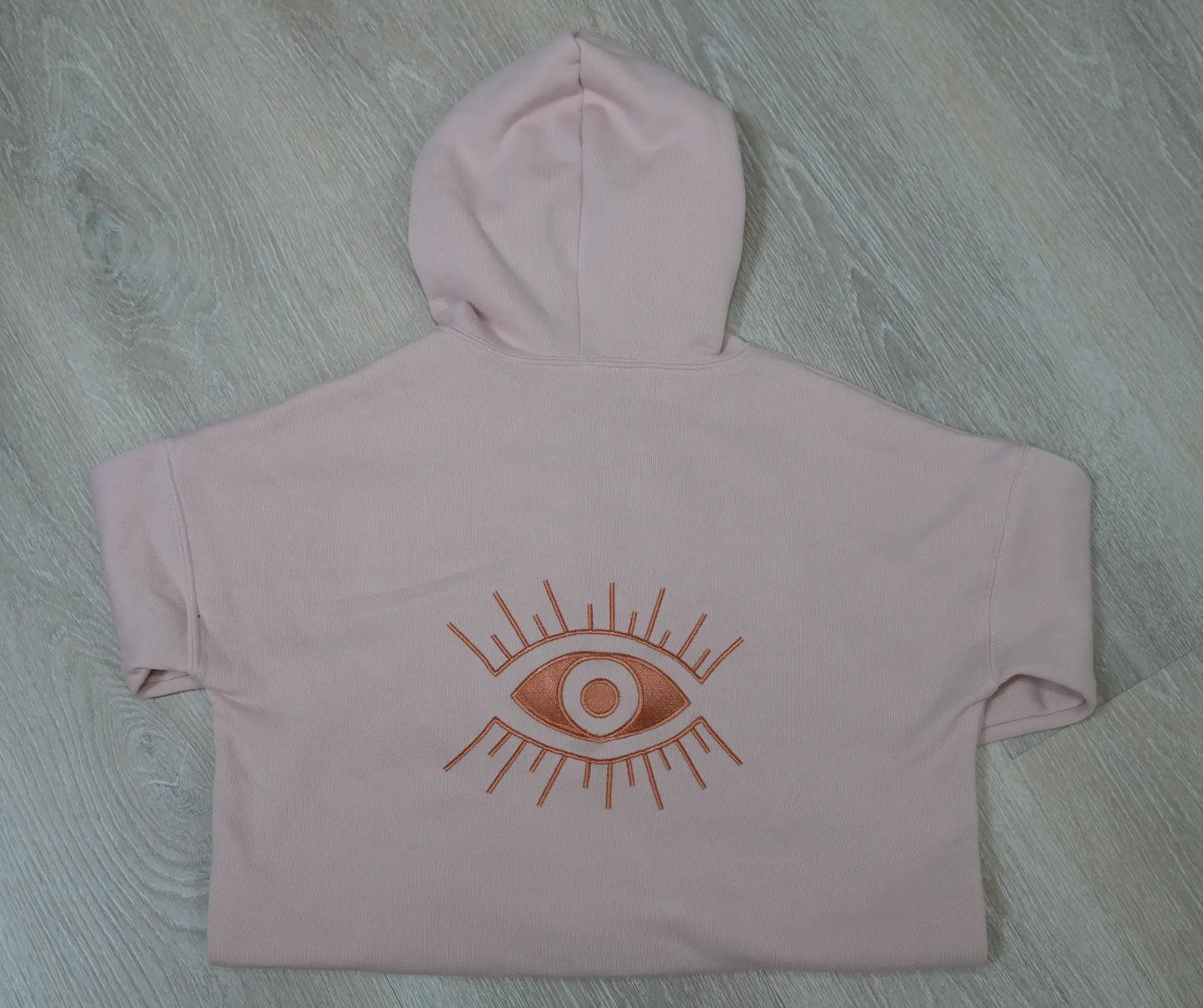 A pink hoodie with a embroidered Evil Eye graphic on the back, displayed on a wooden surface.