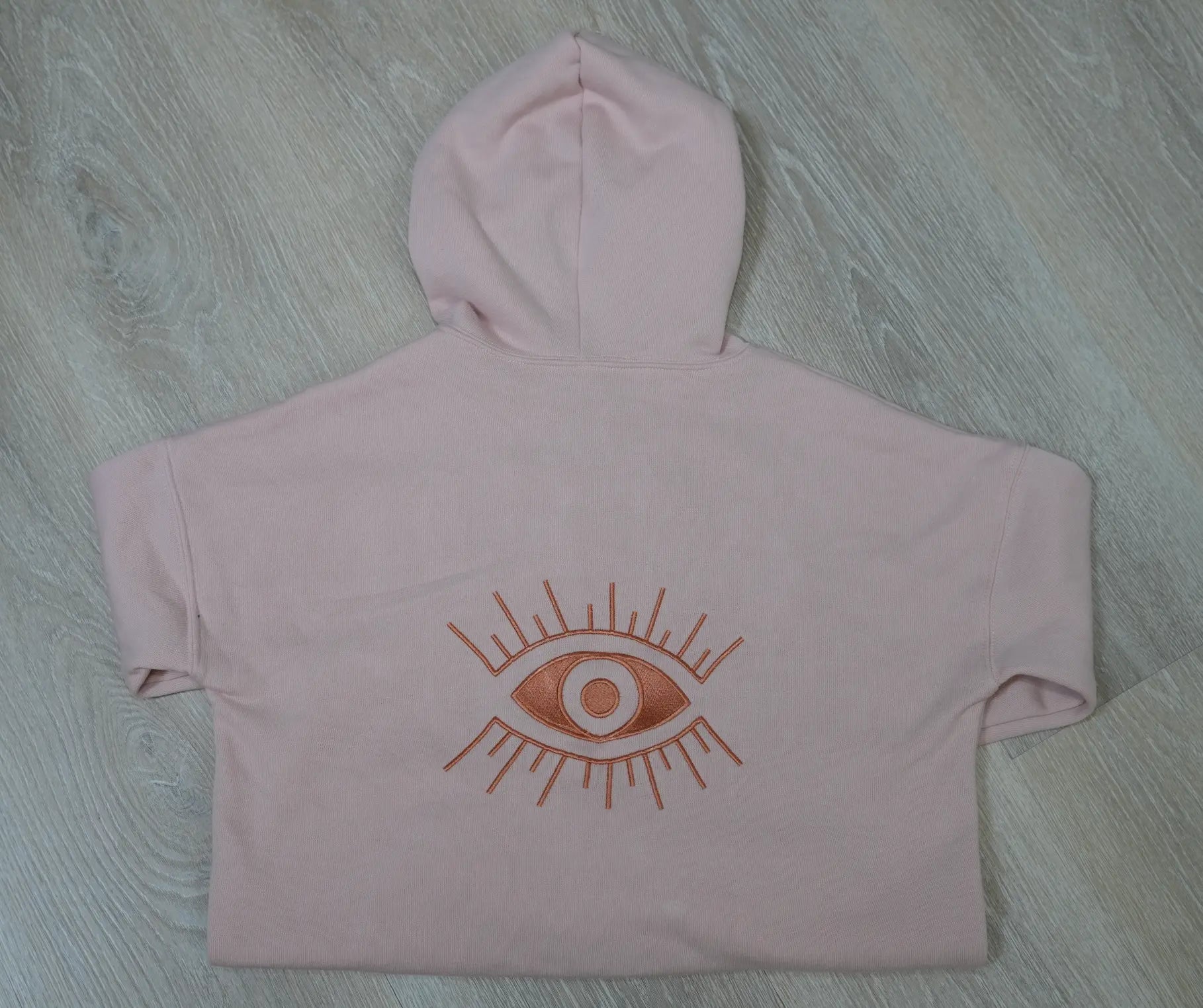 A pink hoodie with a embroidered Evil Eye graphic on the back, displayed on a wooden surface.