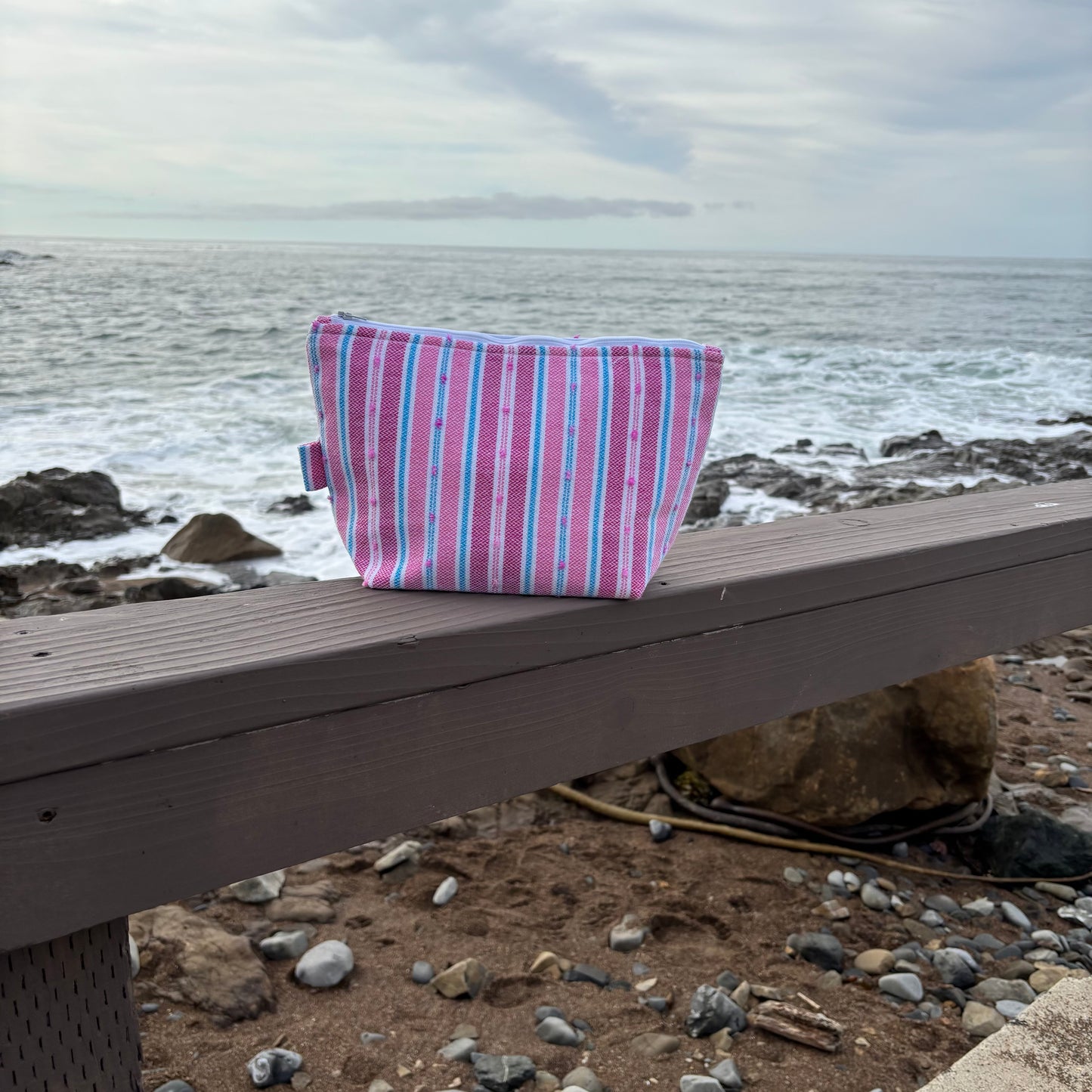 A large striped flamingo-colored makeup bag placed on a wooden surface with a beach background.