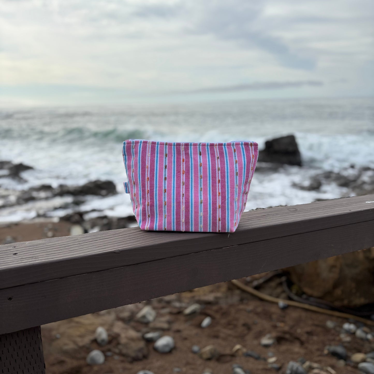 A medium sized striped flamingo-colored makeup bag placed on a wooden surface with a beach background.
