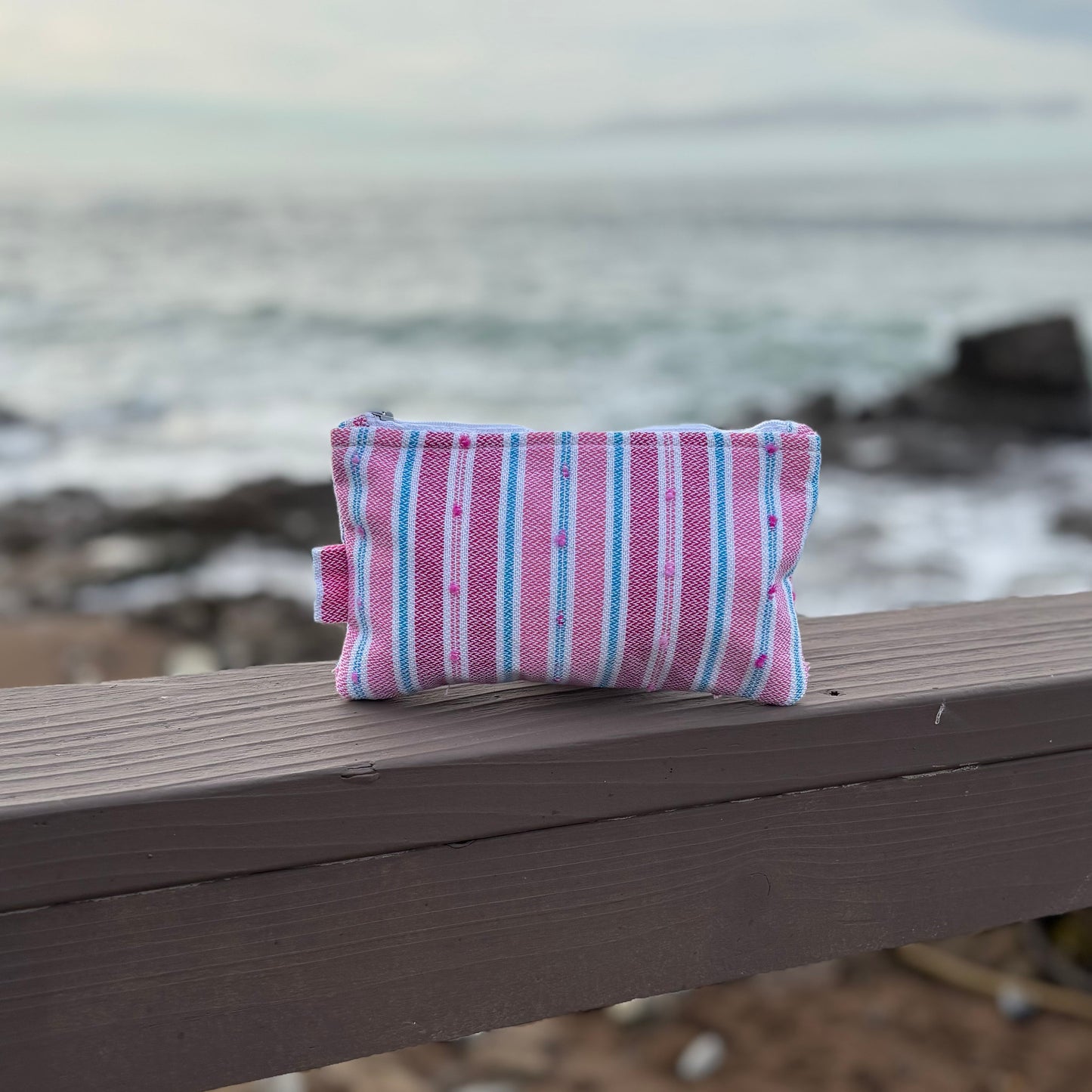 A small striped flamingo-colored makeup bag placed on a wooden surface with a beach background.