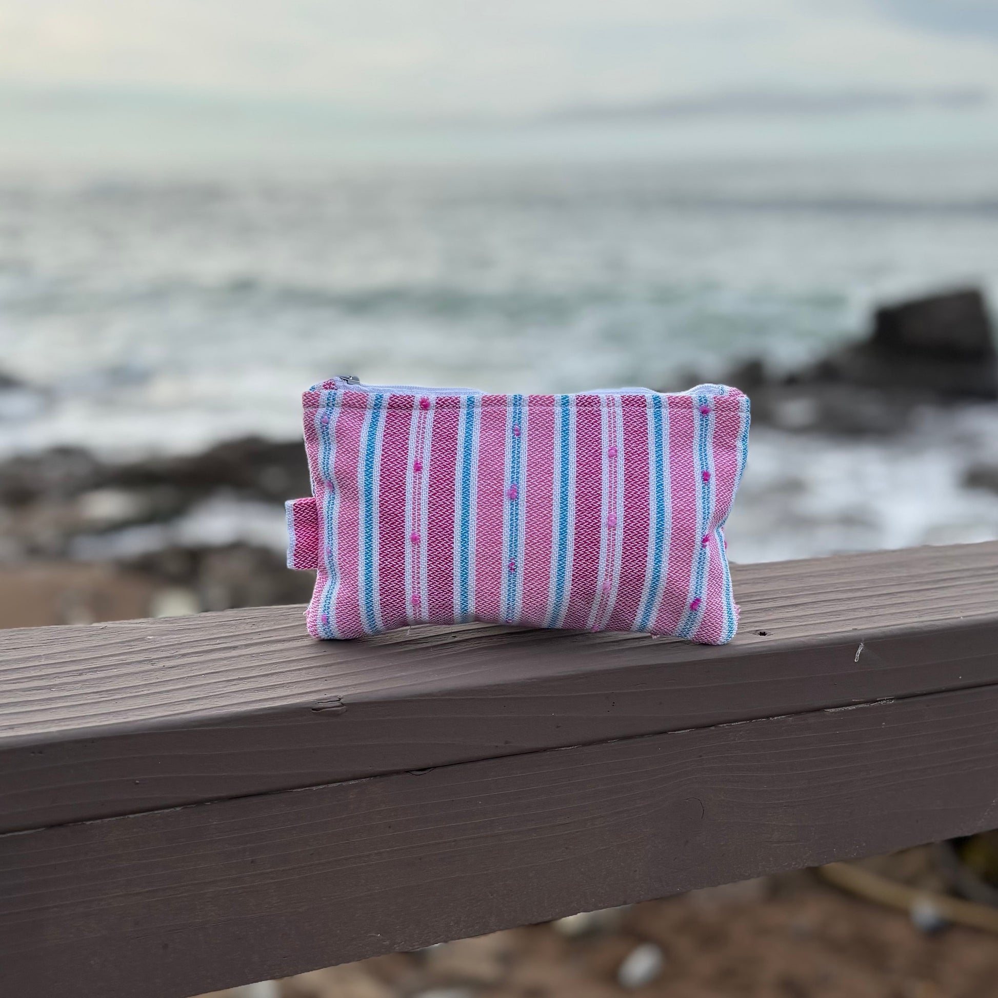 A small striped flamingo-colored makeup bag placed on a wooden surface with a beach background.