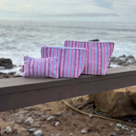 Three striped flamingo-colored makeup bags of different sizes placed on a wooden surface with a beach background.