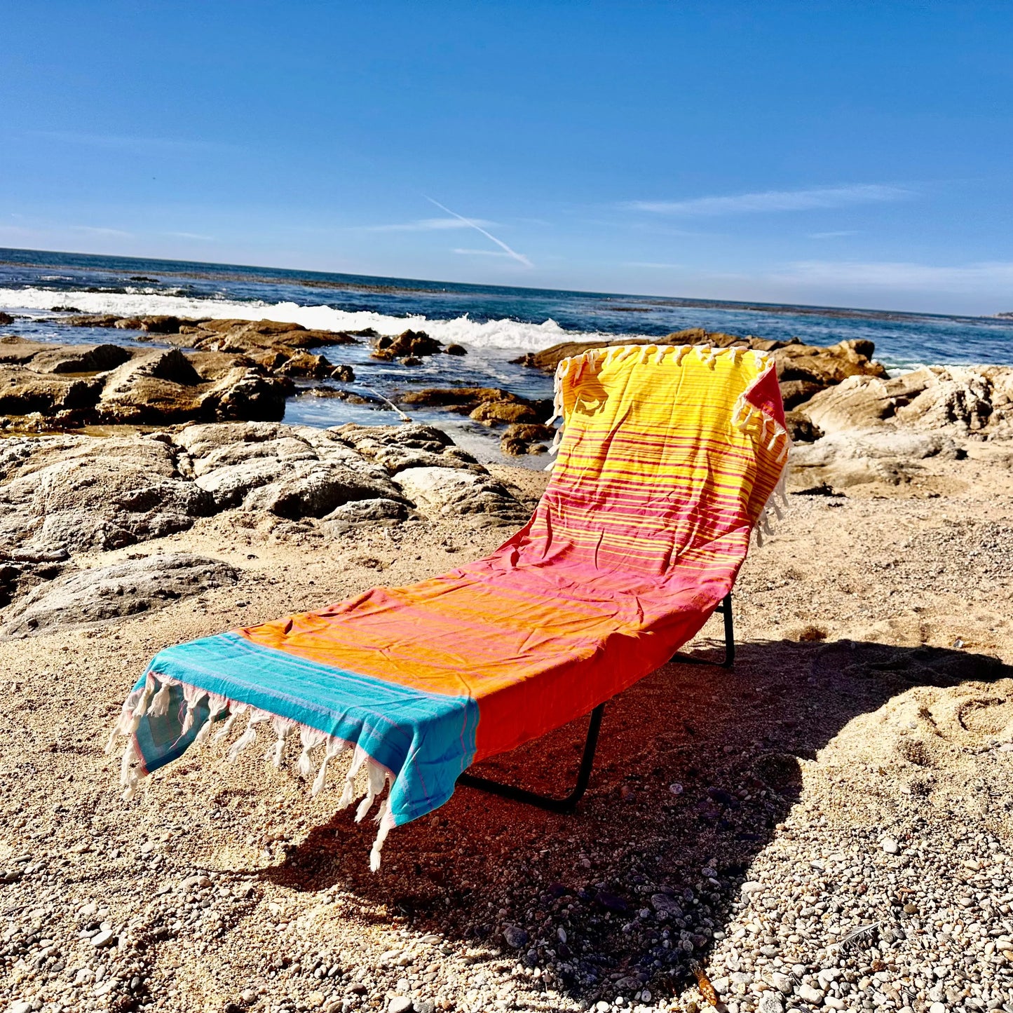 Colorful towel draped over a beach chair on a rocky beach with ocean waves in the background.