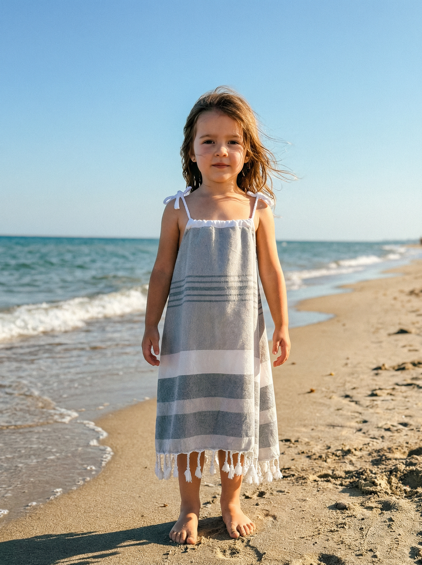 Young girl in a grey and white striped dress with white tassels and white tied shoulder straps.  Standing on a beach with ocean waves in the background