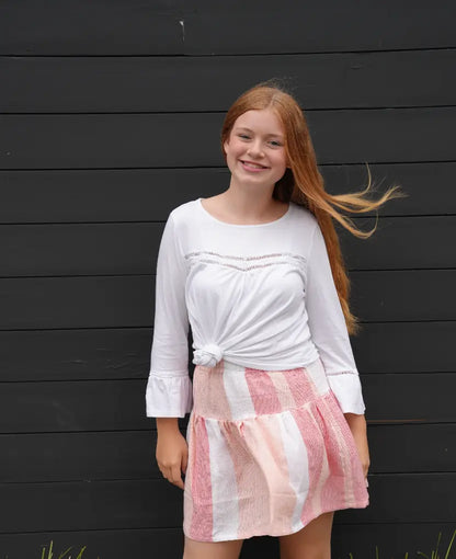 Woman wearing a white top and peach and white striped skirt against a black wooden wall.