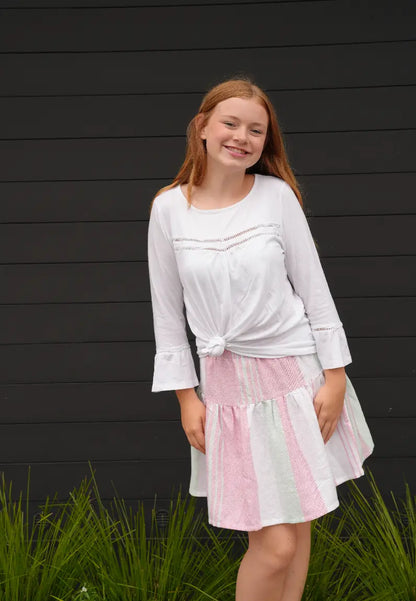 Young girl wearing a white top and pink, green and white striped skirt standing against a black wall with green grass at the bottom.