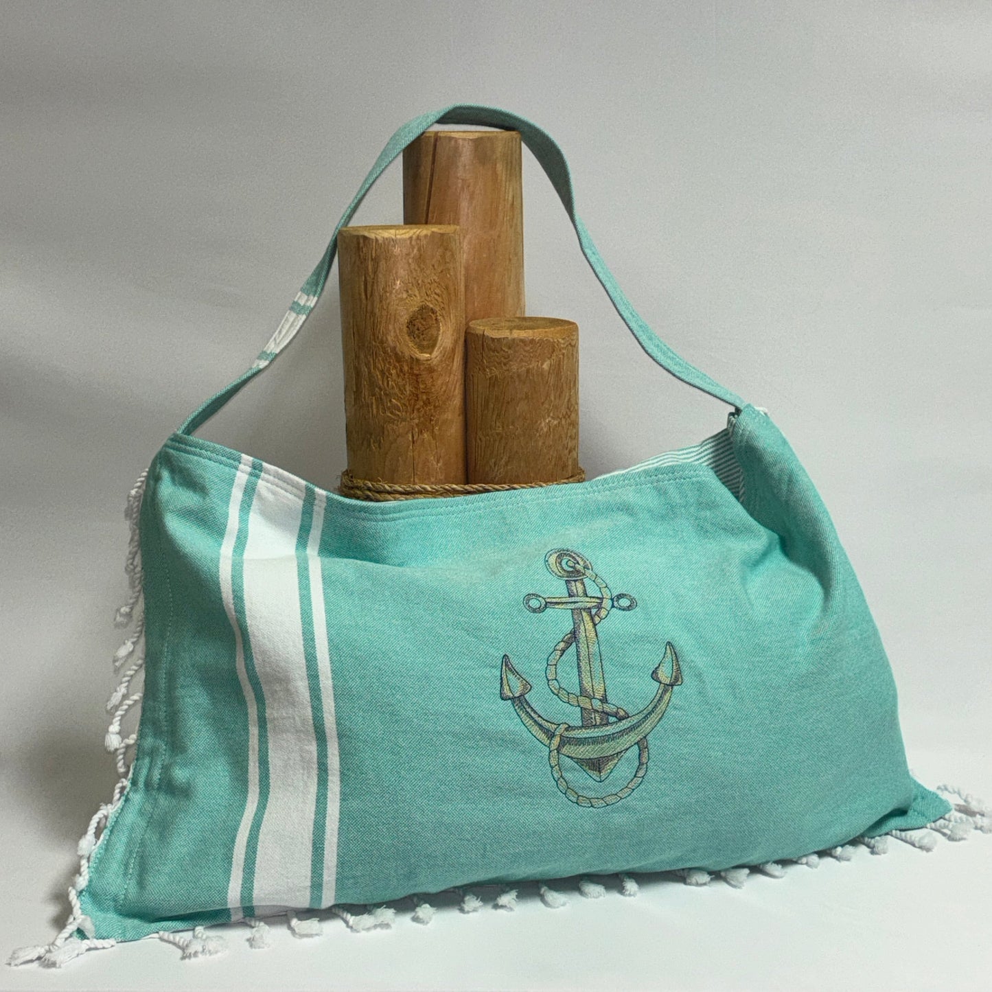 A mint green canvas shoulder bag with white stripes, a white tassel fringe, and a navy anchor graphic on the front. The bag is styled with three wooden cylindrical props against a neutral light gray background.