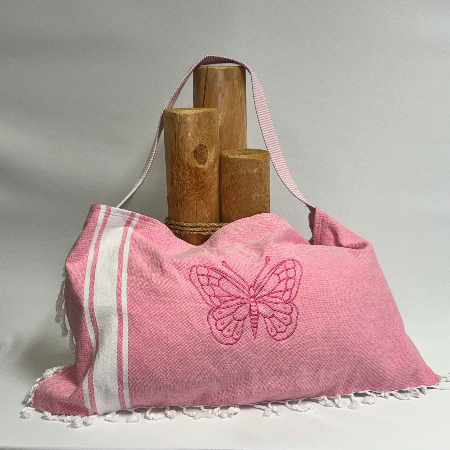 A pink fabric shoulder bag with a white striped side panel, white fringe trim, and an embroidered butterfly motif on the front. The bag is shown with a pink-and-white striped strap, styled with three wooden cylindrical props against a light gray studio background.