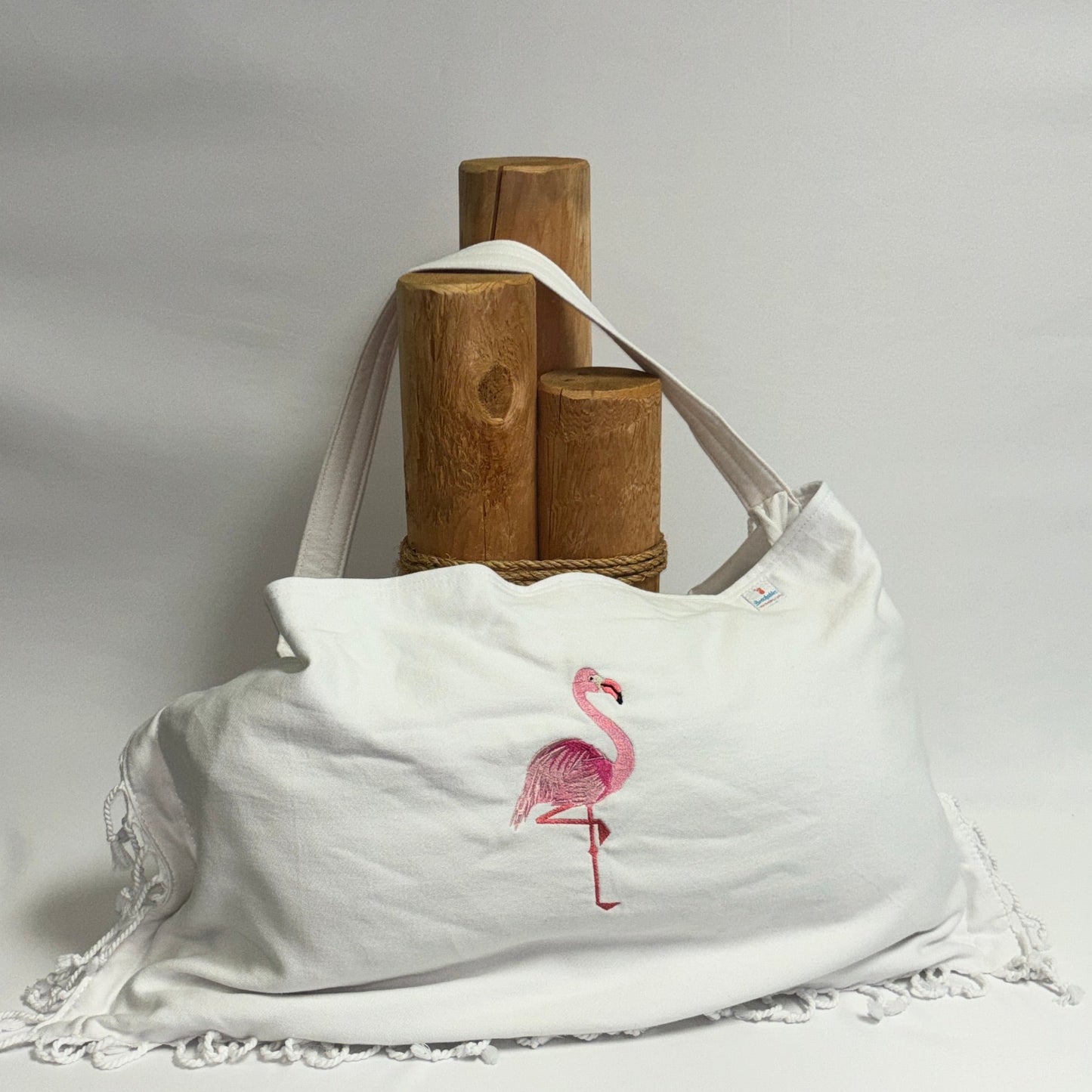 A white canvas shoulder bag with a pink flamingo graphic and white lace trim, displayed on a draped white fabric backdrop with wooden display stands behind it.