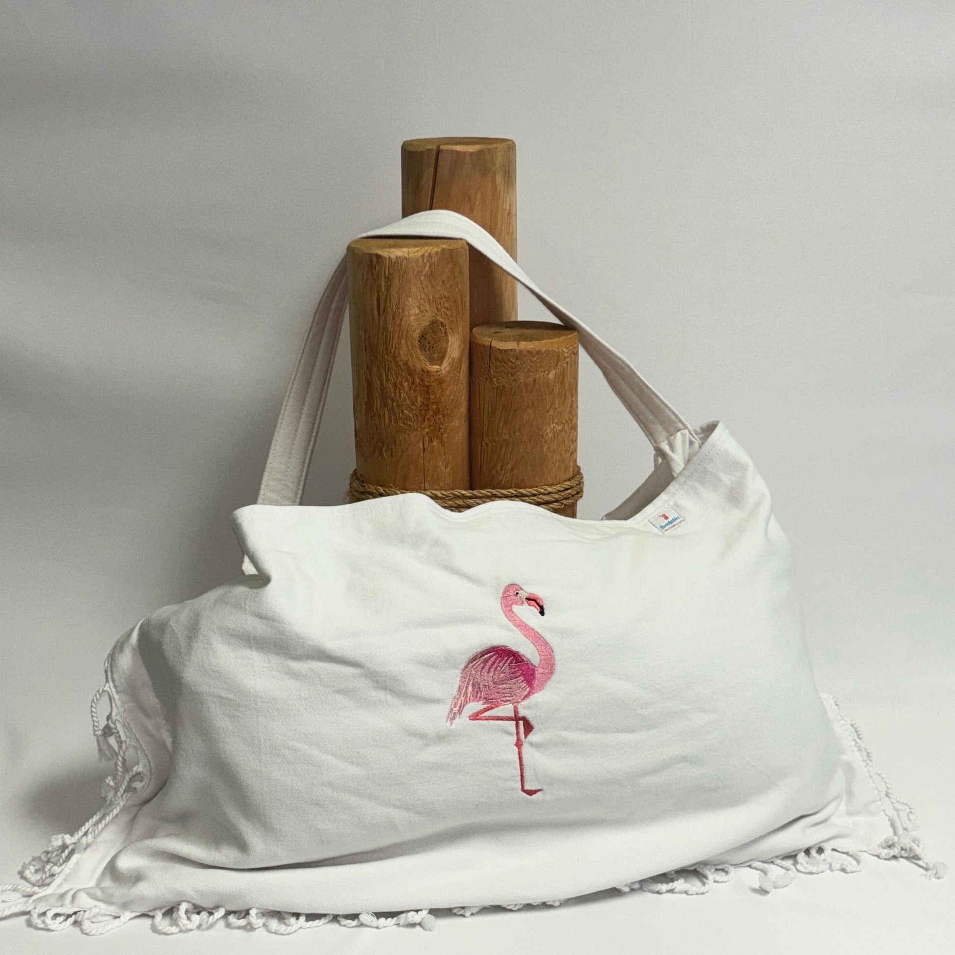 A white canvas shoulder bag with a pink flamingo graphic and white lace trim, displayed on a draped white fabric backdrop with wooden display stands behind it.