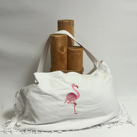 A white canvas shoulder bag with a pink flamingo graphic and white lace trim, displayed on a draped white fabric backdrop with wooden display stands behind it.