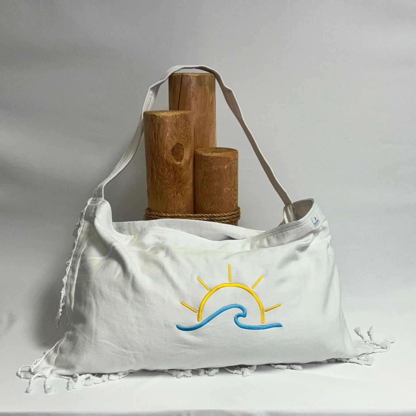 A white canvas shoulder bag with fringe details and a yellow and blue sun-and-wave graphic on the front, displayed on a draped white tablecloth with wooden props behind it.