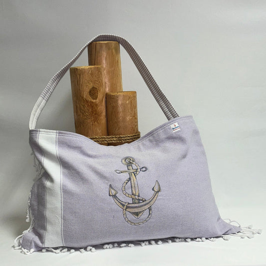 A lavender and white canvas shoulder bag with a white tassel fringe, featuring an embroidered anchor design on the front. The bag is displayed on a draped white fabric table with three wooden display stands behind it.