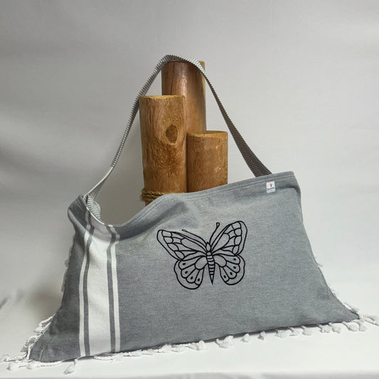 A gray fabric shoulder bag with a black embroidered butterfly design, resting on a draped white tablecloth with wooden display stands behind it.