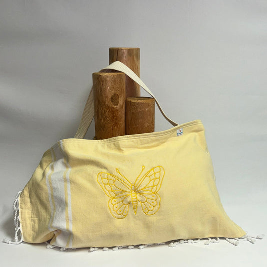A pale yellow fabric shoulder bag with a yellow embroidered butterfly design and white fringe, displayed on a draped white fabric surface with wooden display blocks behind it.