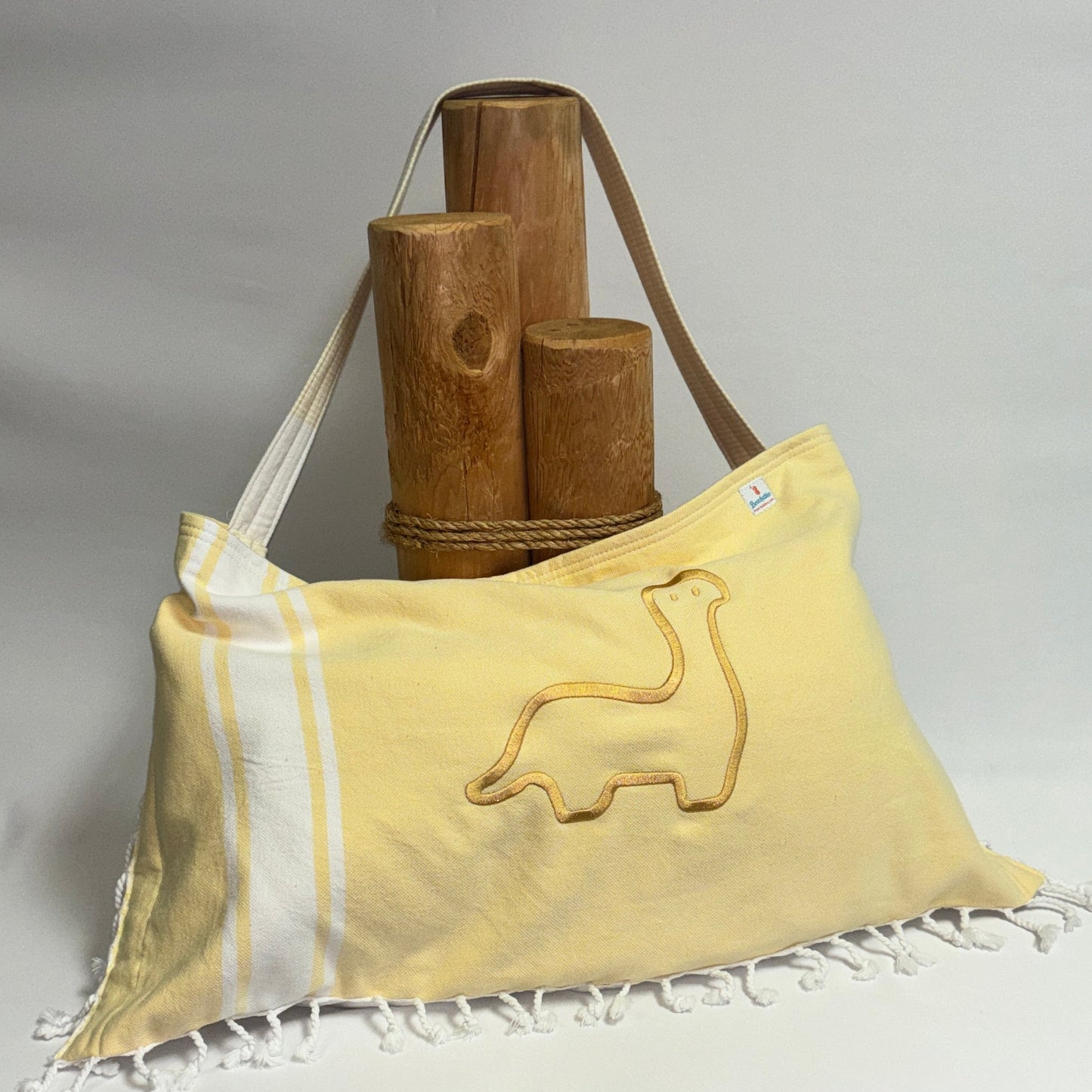 A pale yellow Hebrew towel bag with white fringe, featuring a yellow embroidered dinosaur design and a yellow shoulder strap, displayed on a white draped tablecloth with wooden props in the background.