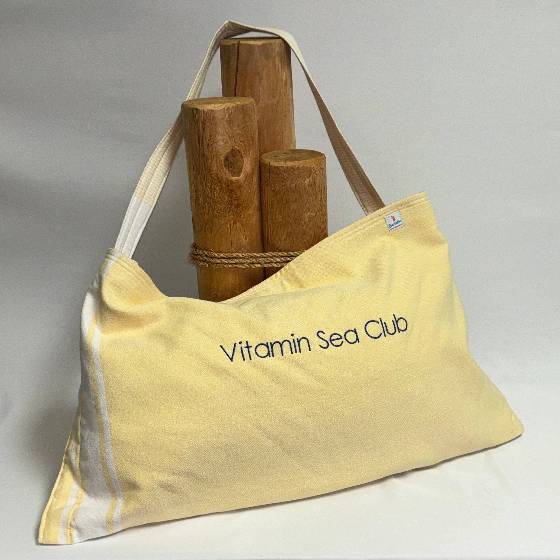 A pale yellow canvas tote bag with 'Vitamin Sea Club' printed in navy blue on the front, displayed on a draped white fabric backdrop next to wooden props.