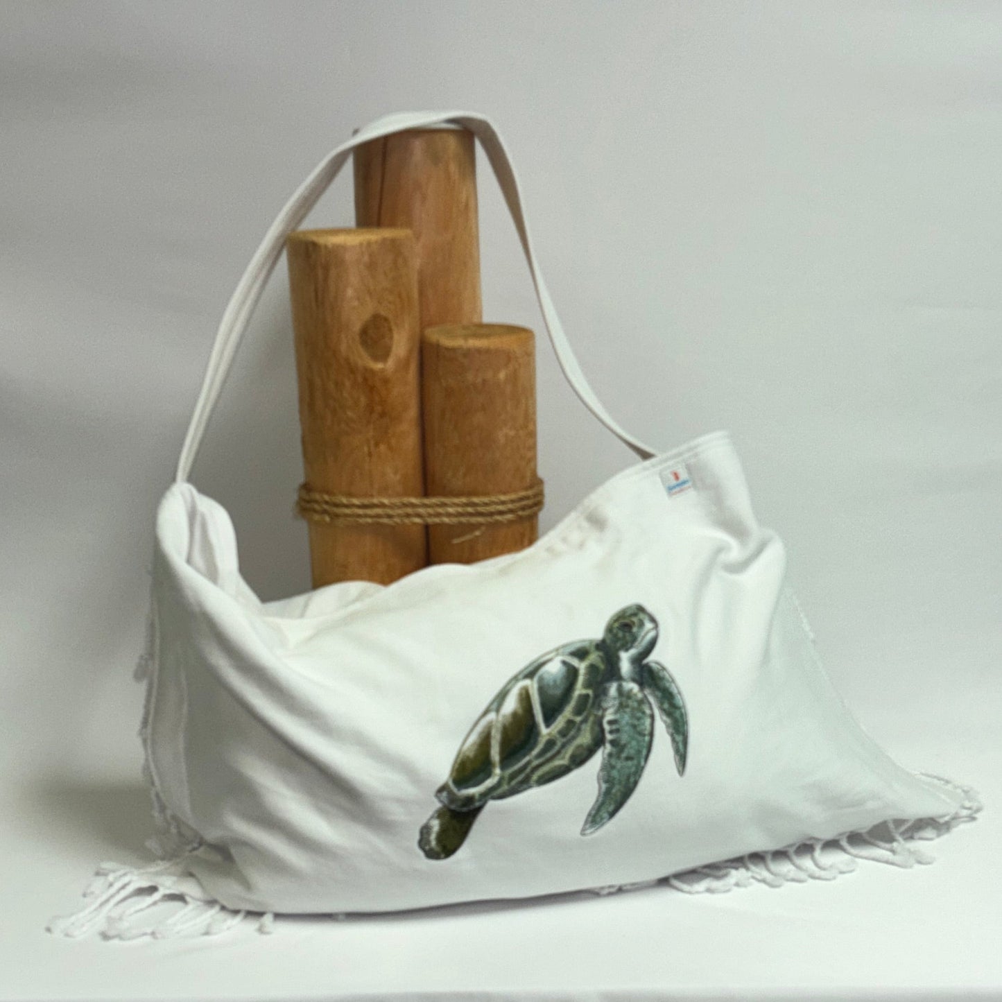 A white canvas shoulder bag with a green sea turtle illustration on the front, displayed on a draped white tablecloth with bamboo props behind it.