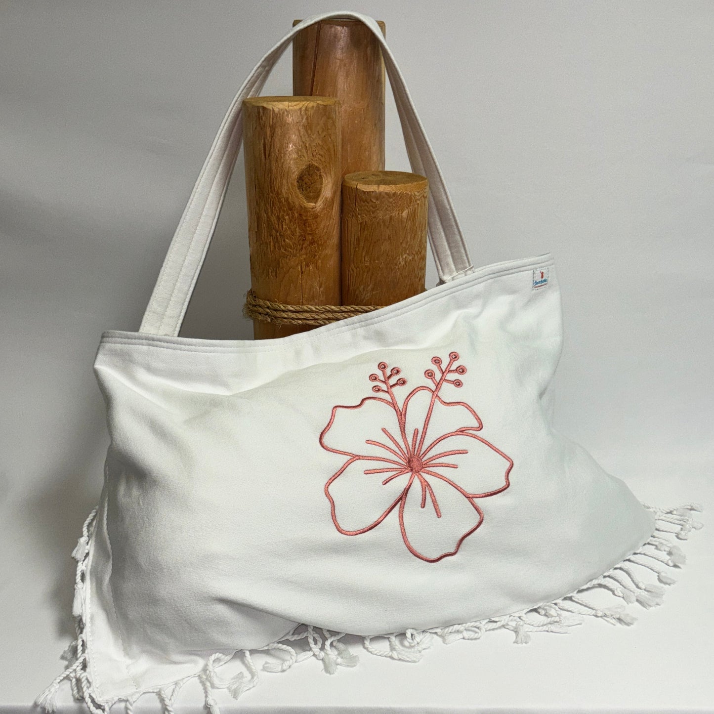 A white canvas tote bag with a rose pink hibiscus flower embroidery on the front, resting on a draped white fabric surface with wooden props behind it.