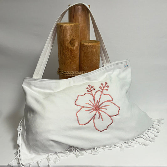 A white canvas tote bag with a rose pink hibiscus flower embroidery on the front, resting on a draped white fabric surface with wooden props behind it.