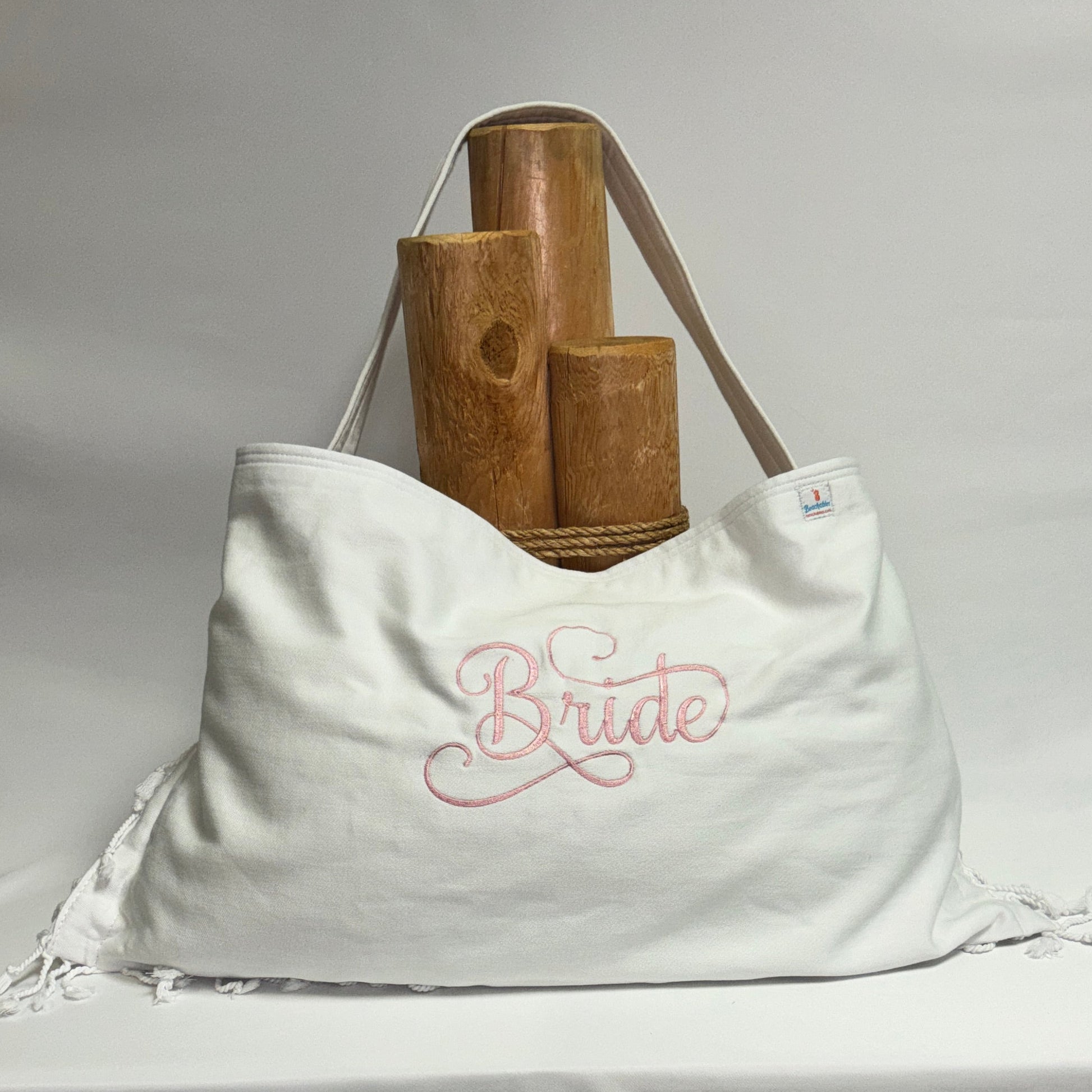 A white fabric tote bag with a pink embroidered script reading 'Bride' on the front, displayed on a draped white tablecloth with wooden props behind it.