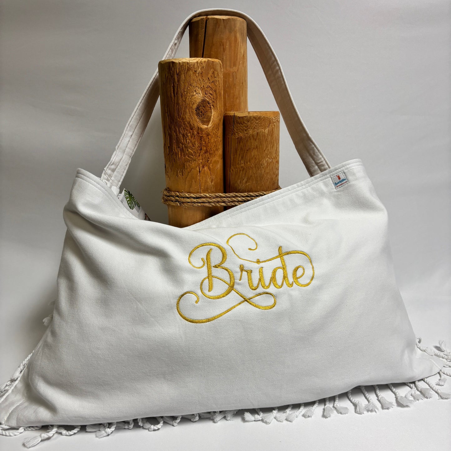 A white canvas tote bag with gold embroidered 'Bride' script on the front, displayed on a white draped surface with white fringe trim. The bag is styled with two wooden cylindrical props and rope handles.