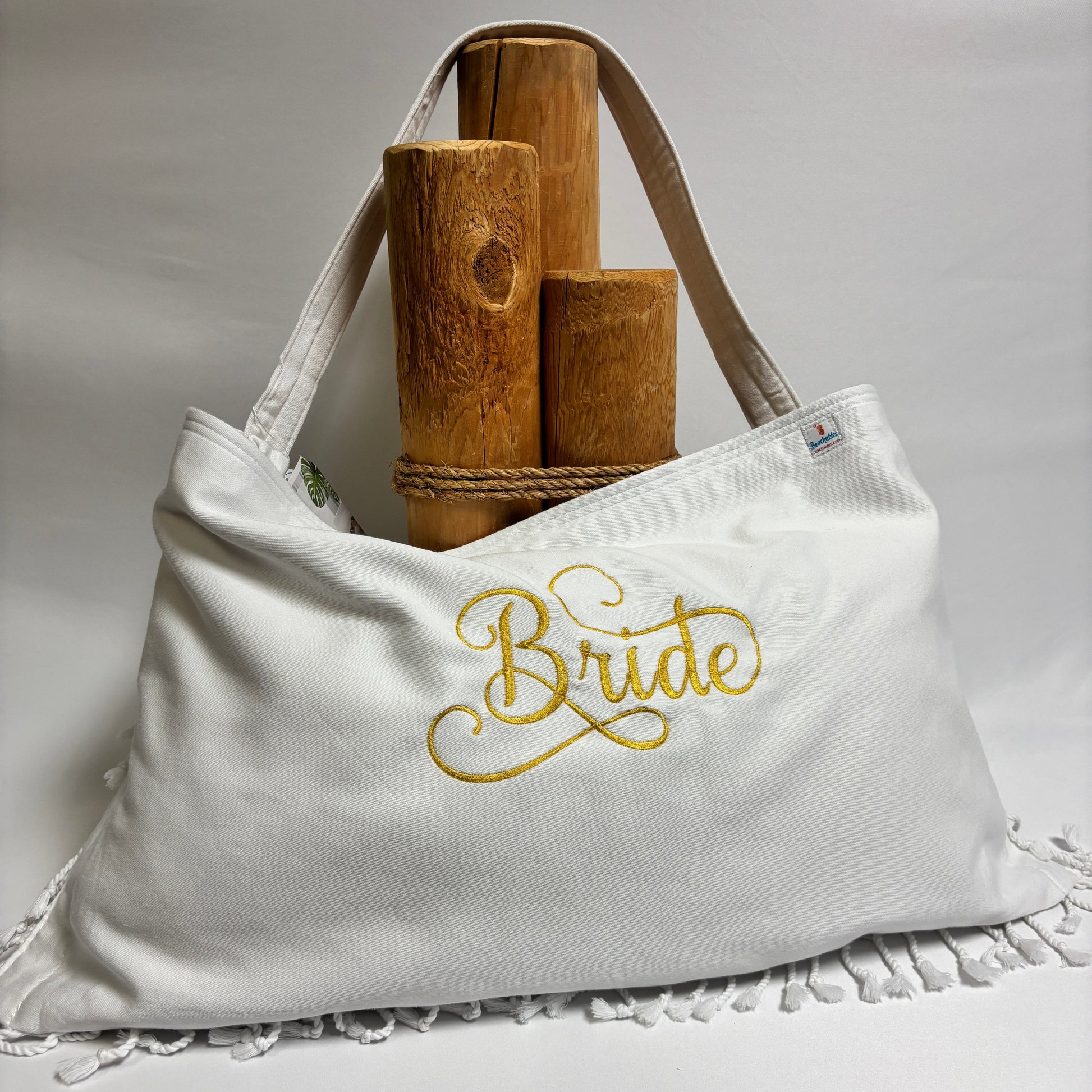 A white canvas tote bag with gold embroidered 'Bride' script on the front, displayed on a white draped surface with white fringe trim. The bag is styled with two wooden cylindrical props and rope handles.