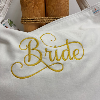 A white canvas tote bag with 'Bride' embroidered in yellow-gold script, displayed on a rustic wooden stand with a rope band.