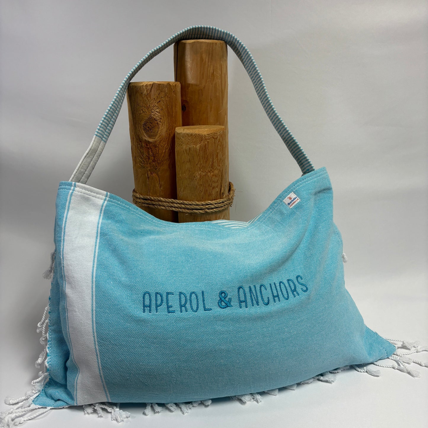 A light blue and white fringed shoulder bag with 'APEROL & ANCHORS' printed on the front, displayed on a white surface with wooden props against a white backdrop.