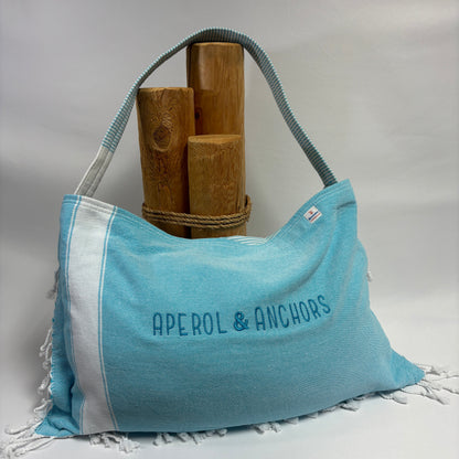 A light blue and white fringed shoulder bag with 'APEROL & ANCHORS' printed on the front, displayed on a white surface with wooden props against a white backdrop.