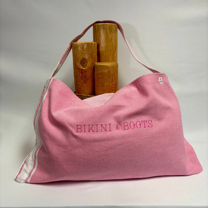 A pink fabric tote bag with 'BIKINI & BOOTS' embroidered on the front, featuring white side stripes and a pink shoulder strap, displayed with three wooden props against a white backdrop.