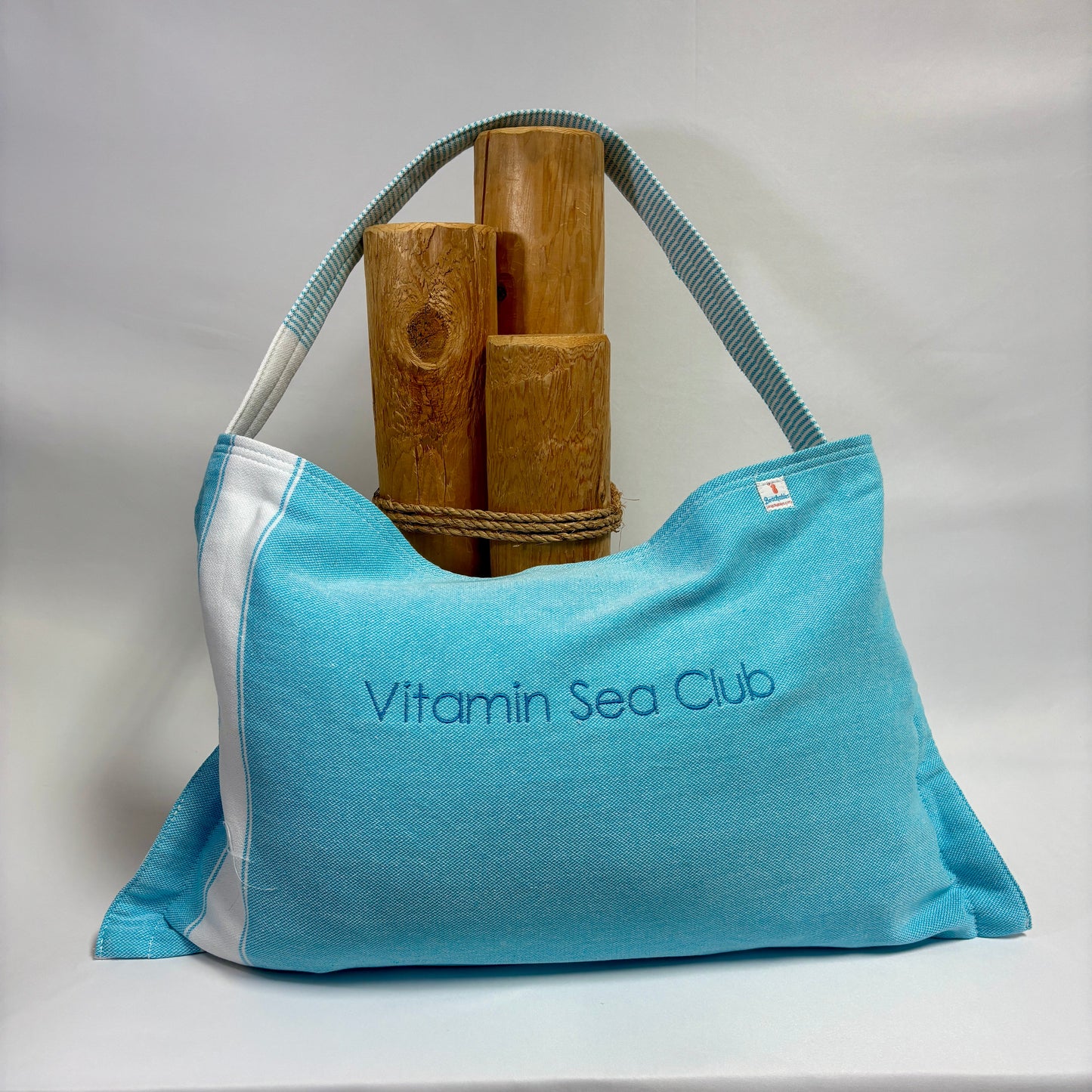 A light blue fabric tote bag with white side panels and a blue-gray shoulder strap, featuring the text 'Vitamin Sea Club' printed on the front. The bag is styled with bamboo poles tied around its upper area, set against a draped white fabric backdrop.