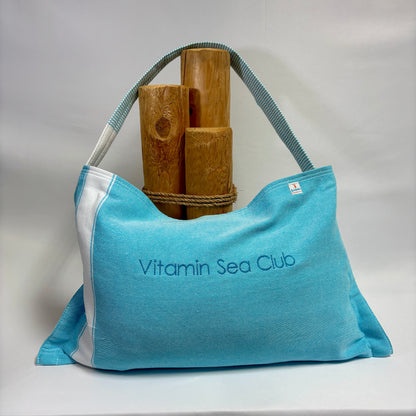 A light blue fabric tote bag with white side panels and a blue-gray shoulder strap, featuring the text 'Vitamin Sea Club' printed on the front. The bag is styled with bamboo poles tied around its upper area, set against a draped white fabric backdrop.