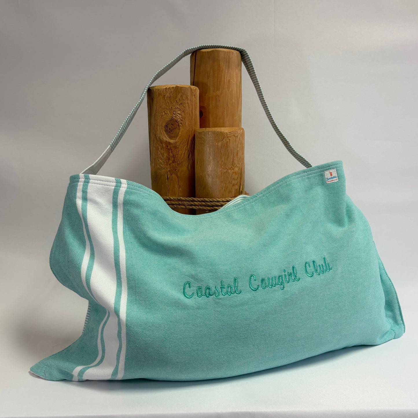 A turquoise and white striped beach towel tote bag with 'Coastal Cougars Club' embroidered on the front, displayed on a white draped fabric background with three wooden blocks behind it.