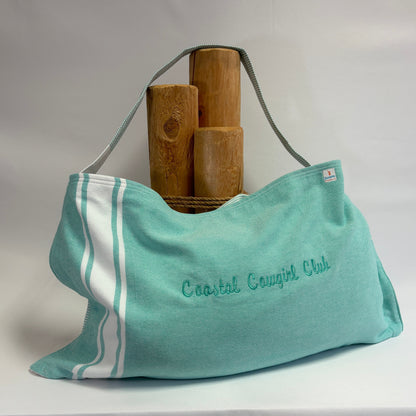 A turquoise and white striped beach towel tote bag with 'Coastal Cougars Club' embroidered on the front, displayed on a white draped fabric background with three wooden blocks behind it.