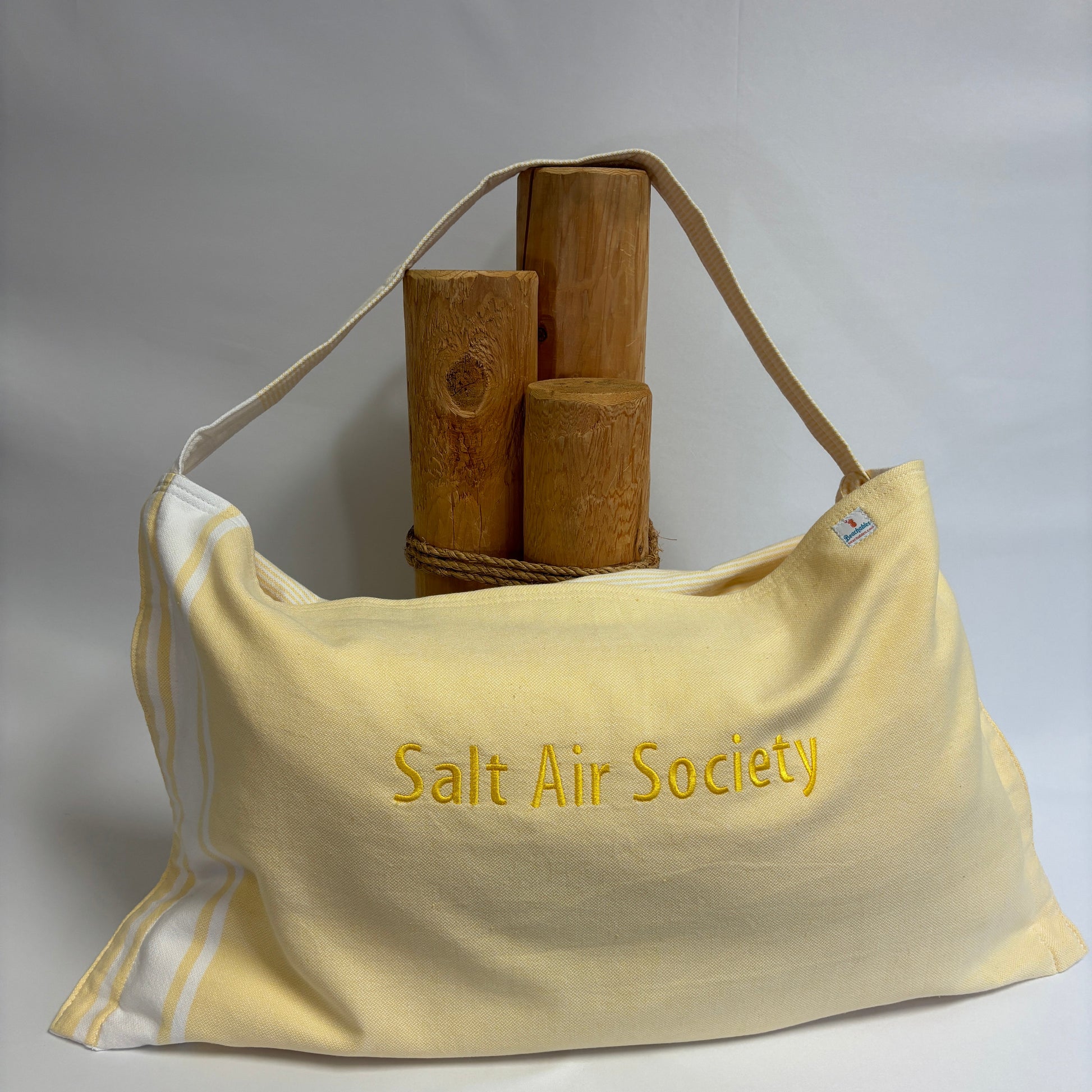 A pale yellow canvas shoulder bag with 'Salt Air Society' embroidered in yellow on the front, featuring a tan shoulder strap and white piping along the side. The bag is styled with three wooden mallets on a white surface against a light gray background.
