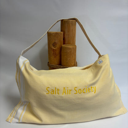 A pale yellow canvas shoulder bag with 'Salt Air Society' embroidered in yellow on the front, featuring a tan shoulder strap and white piping along the side. The bag is styled with three wooden mallets on a white surface against a light gray background.