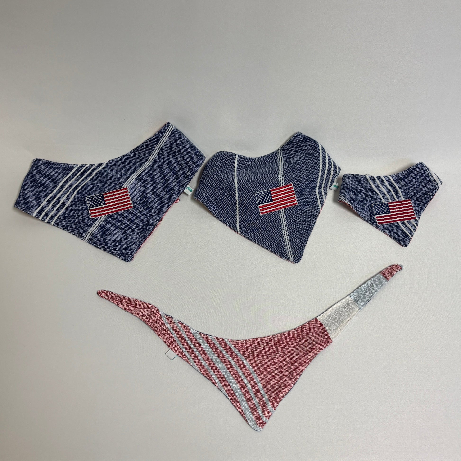 Three dog bandanas laid out on a white surface: two navy blue bandanas with white stripes and American flag patches, and one red bandana with white stripes.