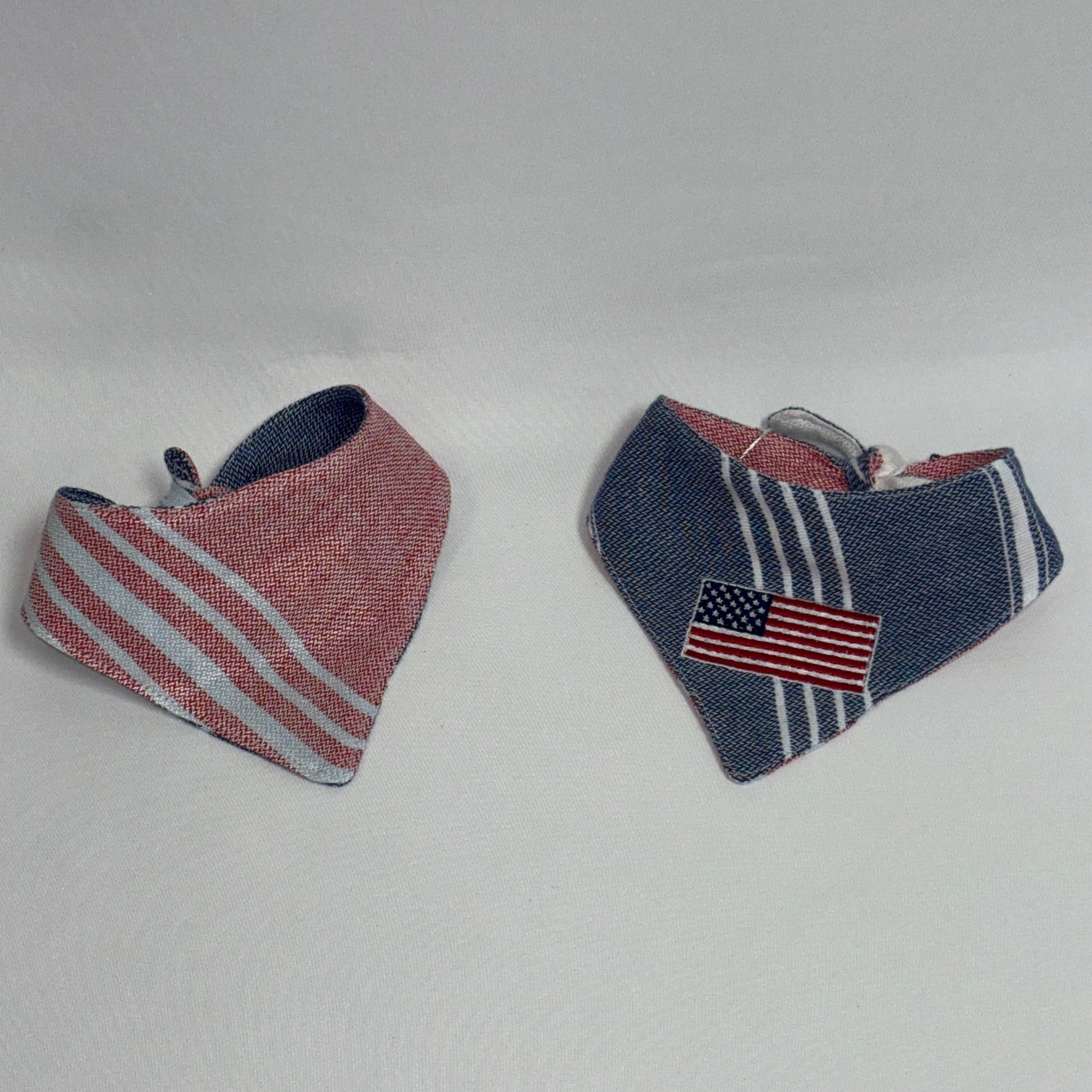 Two small triangular bandanas laid on a light gray background. One is pink with white diagonal stripes, and the other is navy blue with white vertical stripes and a small American flag patch.