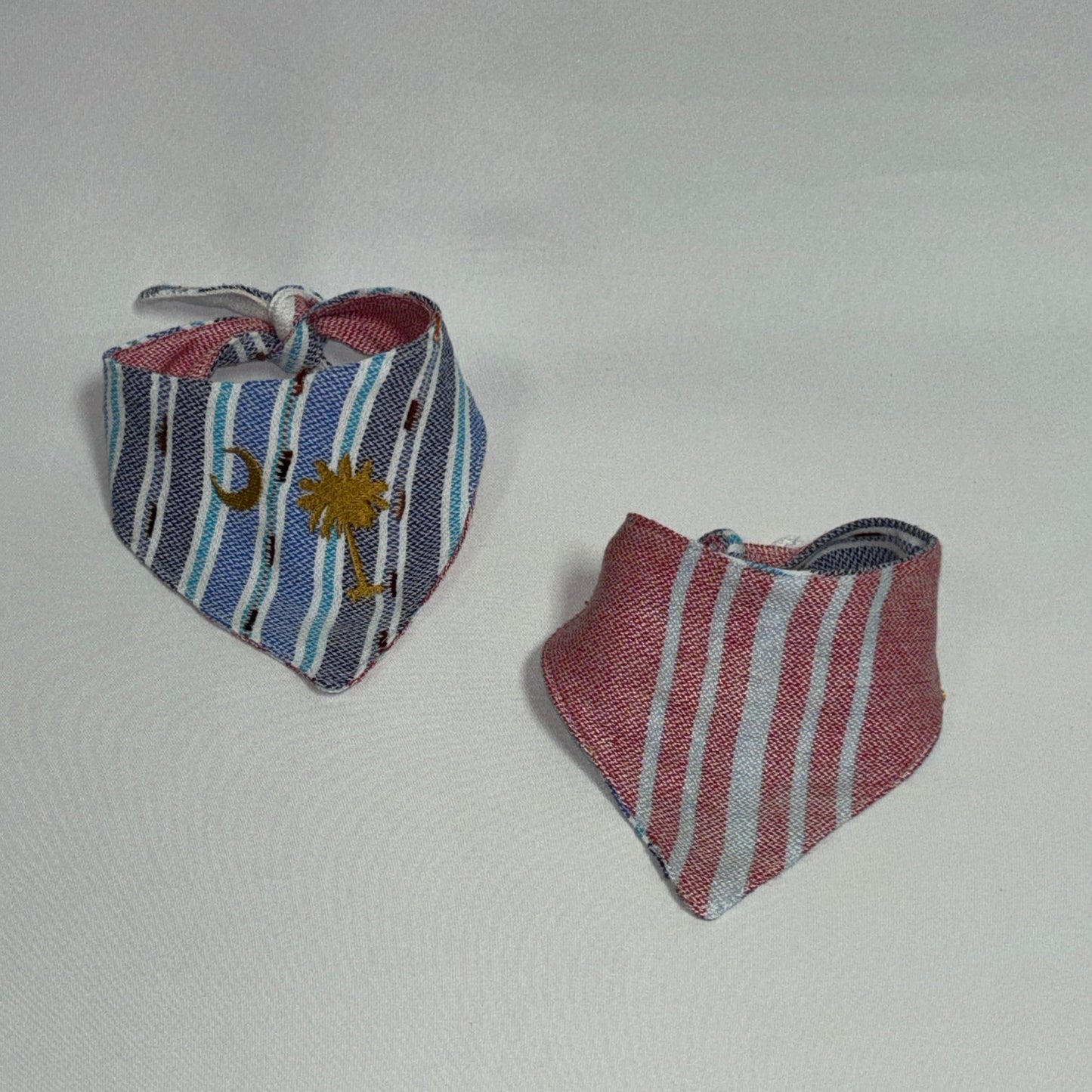 Two small striped fabric bandanas or bib-style accessories on a plain light background. One has blue, white, and gray stripes with a small gold emblem, and the other has pink, white, and gray stripes.