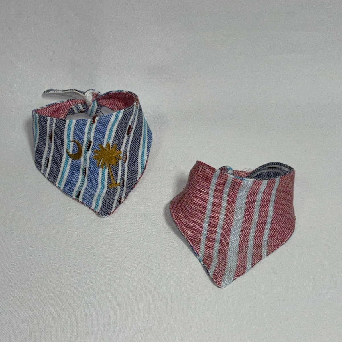 Two small striped fabric bandanas or bib-style accessories on a plain light background. One has blue, white, and gray stripes with a small gold emblem, and the other has pink, white, and gray stripes.