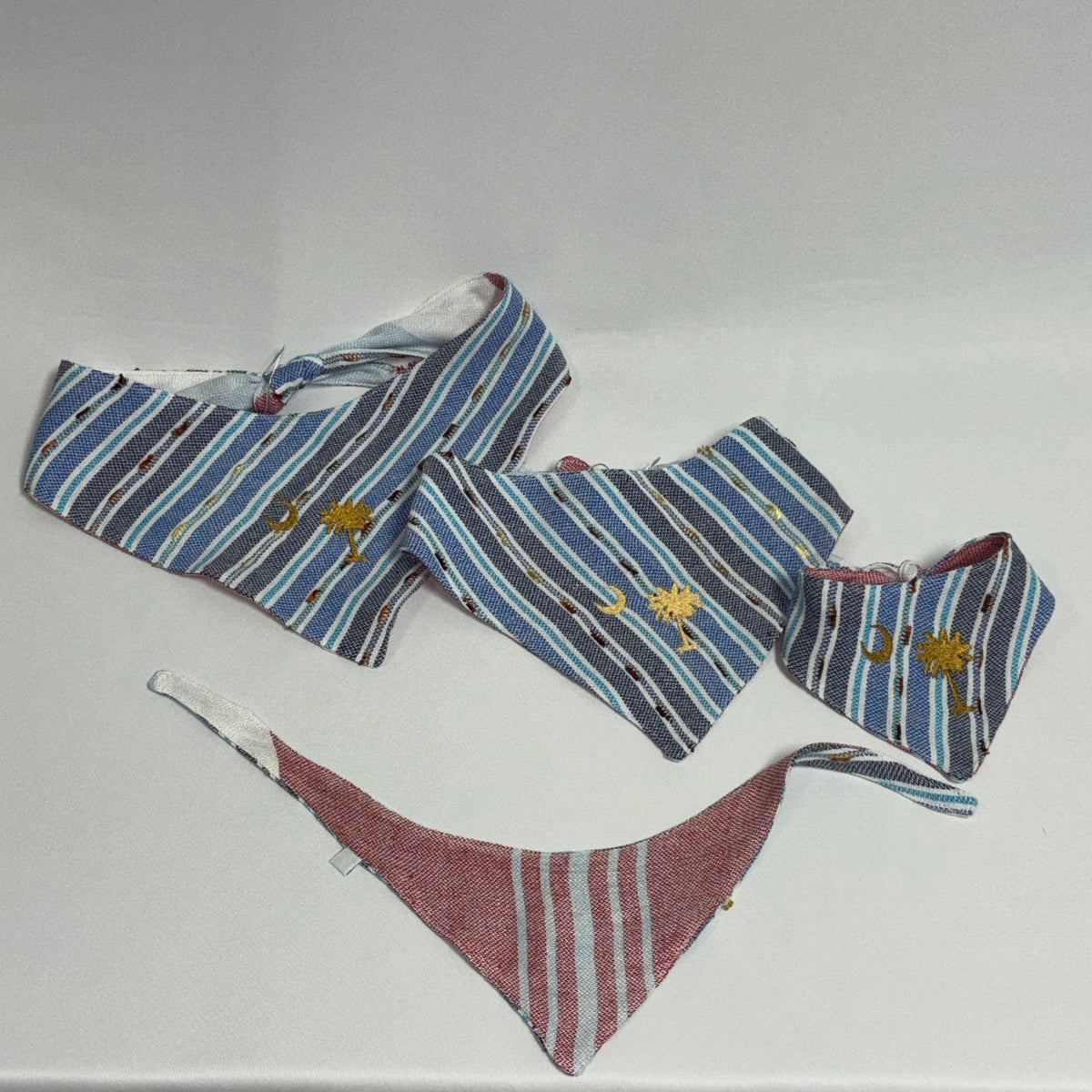 Three striped pet bandanas in blue, red, and white, laid out on a light gray fabric surface. Two are wide triangle bandanas and one is a smaller square bandana, all featuring embroidered yellow flowers and small blue and red details.