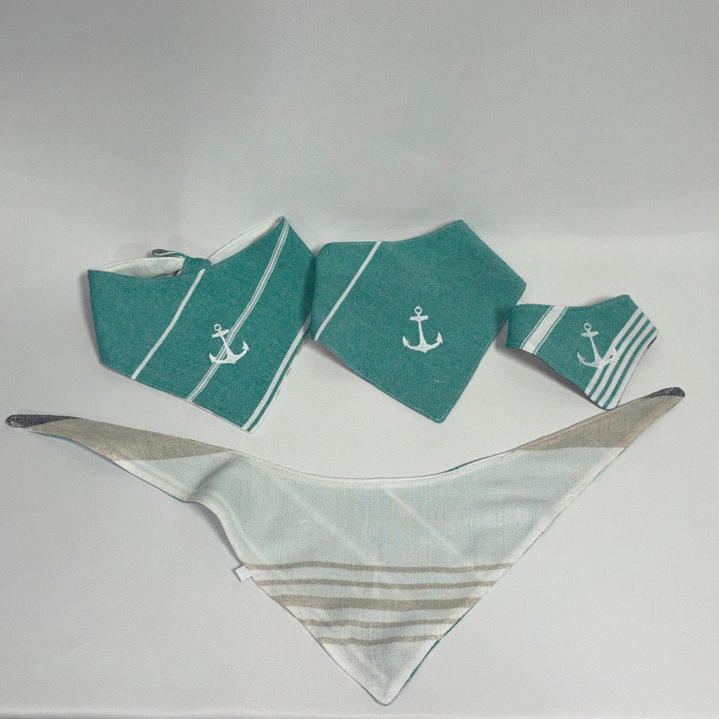 A set of folded pocket squares and a matching pocket square on a white fabric-covered surface. The set includes teal and white striped squares with white anchor motifs.