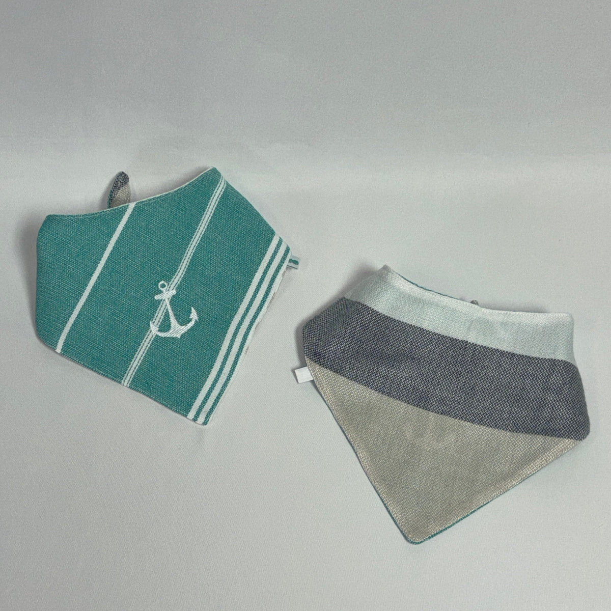 Two fabric baby bibs laid flat on a light gray background. The left bib is teal with white stripes and a white anchor motif.