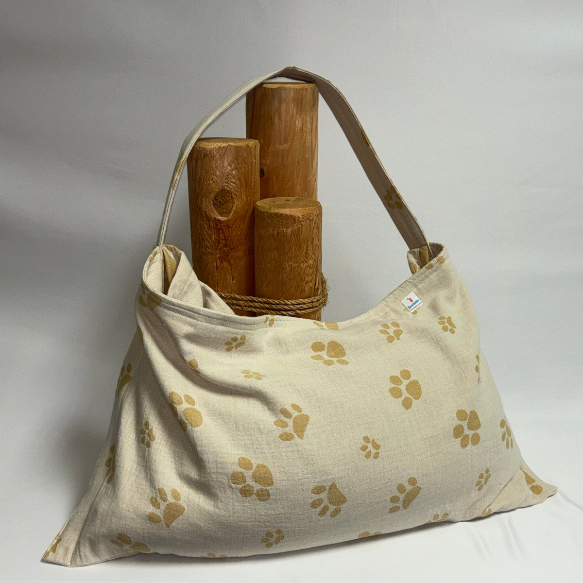 A canvas pet carrier bag in beige with a repeating tan paw print pattern, shown on a draped white fabric backdrop with three wooden dowels inside for support.