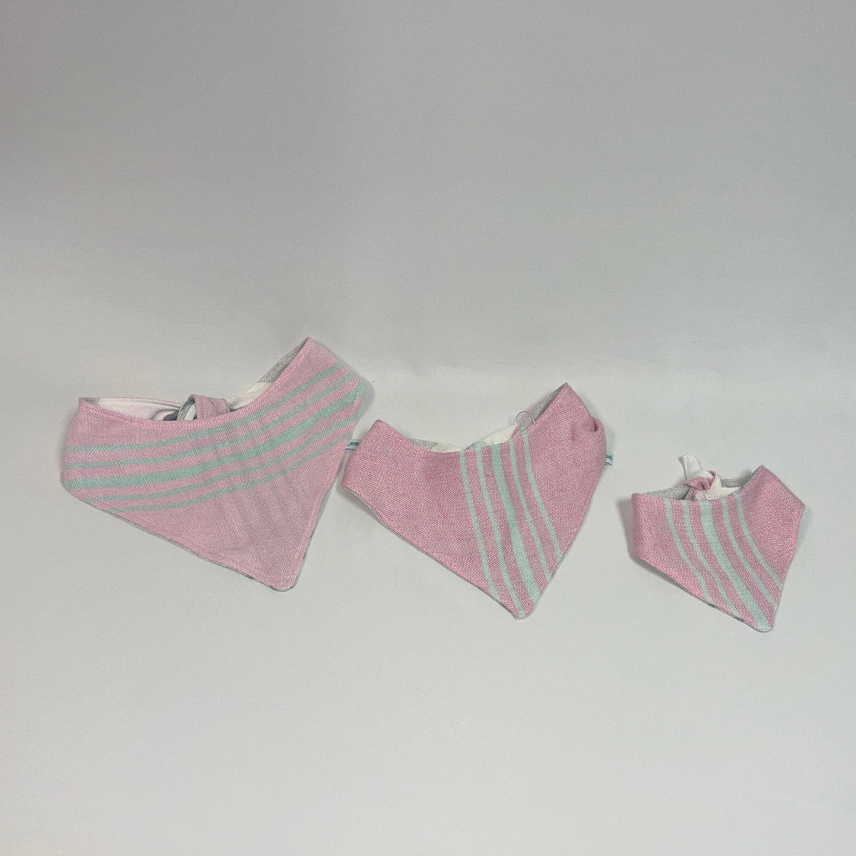 Three pink triangular bandana bibs with light blue stripes in different sizes, arranged horizontally on a plain light gray background.