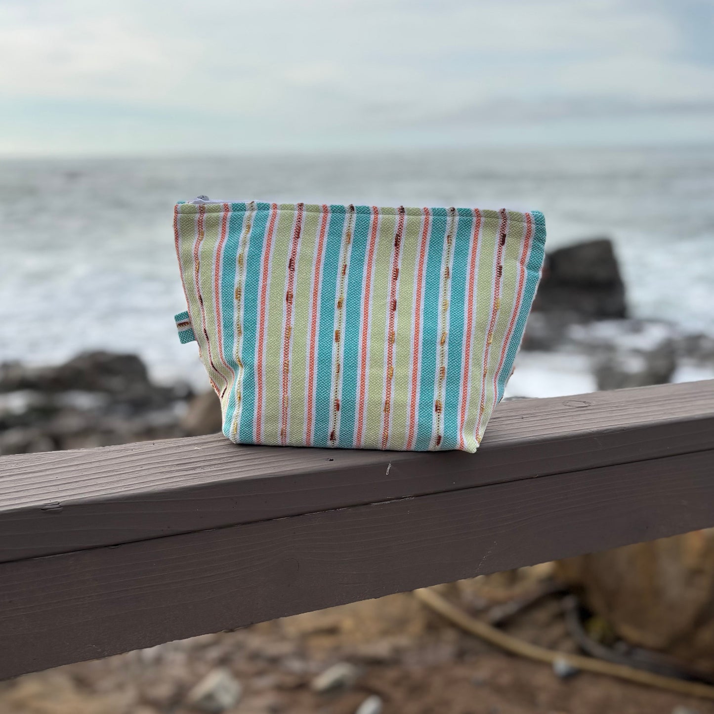 Large sized makeup bag with vibrant  green striped patterns placed on a wooden surface near a coastal backdrop.