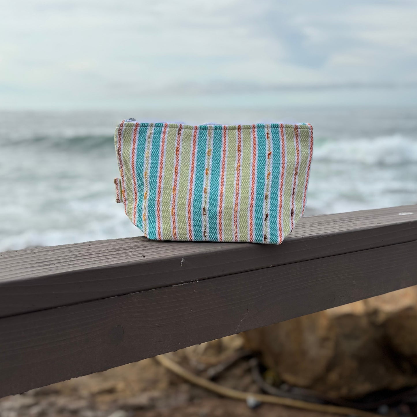 Medium size makeup bag with vibrant  green striped patterns placed on a wooden surface near a coastal backdrop.